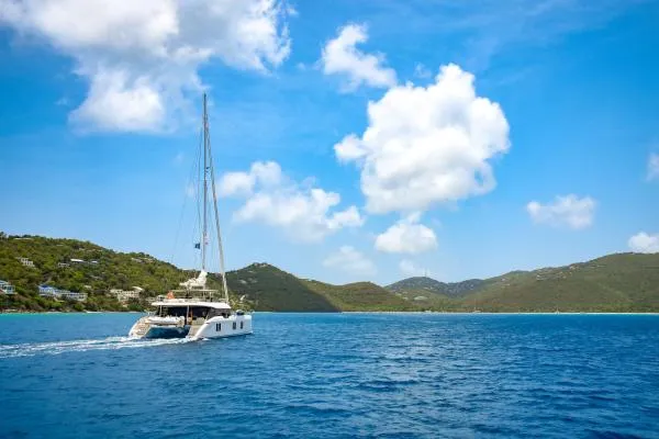 Ocean Vibes Yacht Photos Pics Sunreef 70 yacht sailing in clear blue waters near lush green hills, under a bright sky.