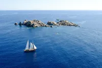 Vintage 1944 Custom Schooner sailing near rocky island in clear blue sea.