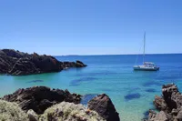 Sailboat Dragonfly 920 on clear blue sea near rocky coastline, under a bright blue sky.