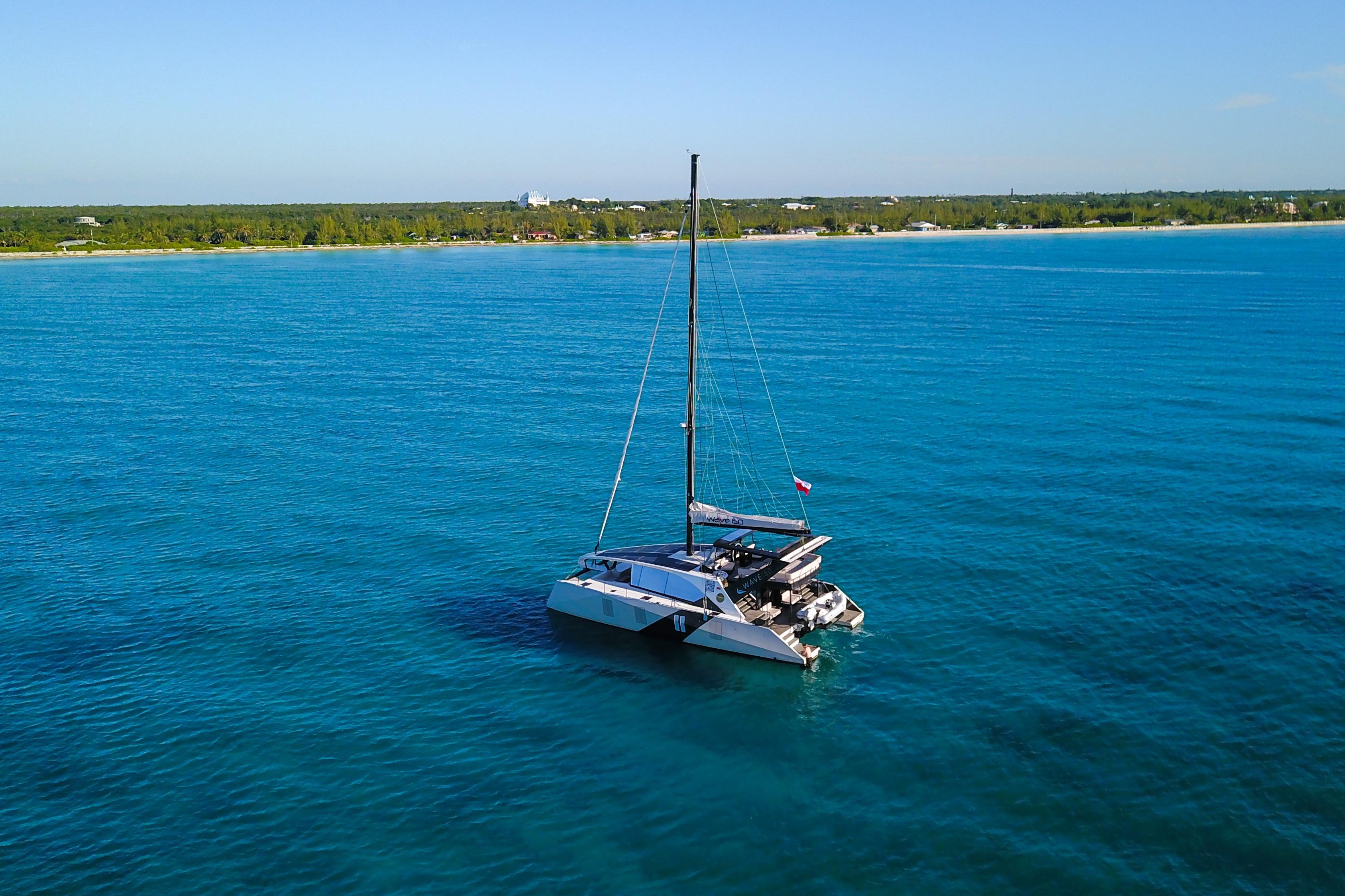 2024 Wave Wave60 catamaran sailing on clear blue ocean near a lush coastline.