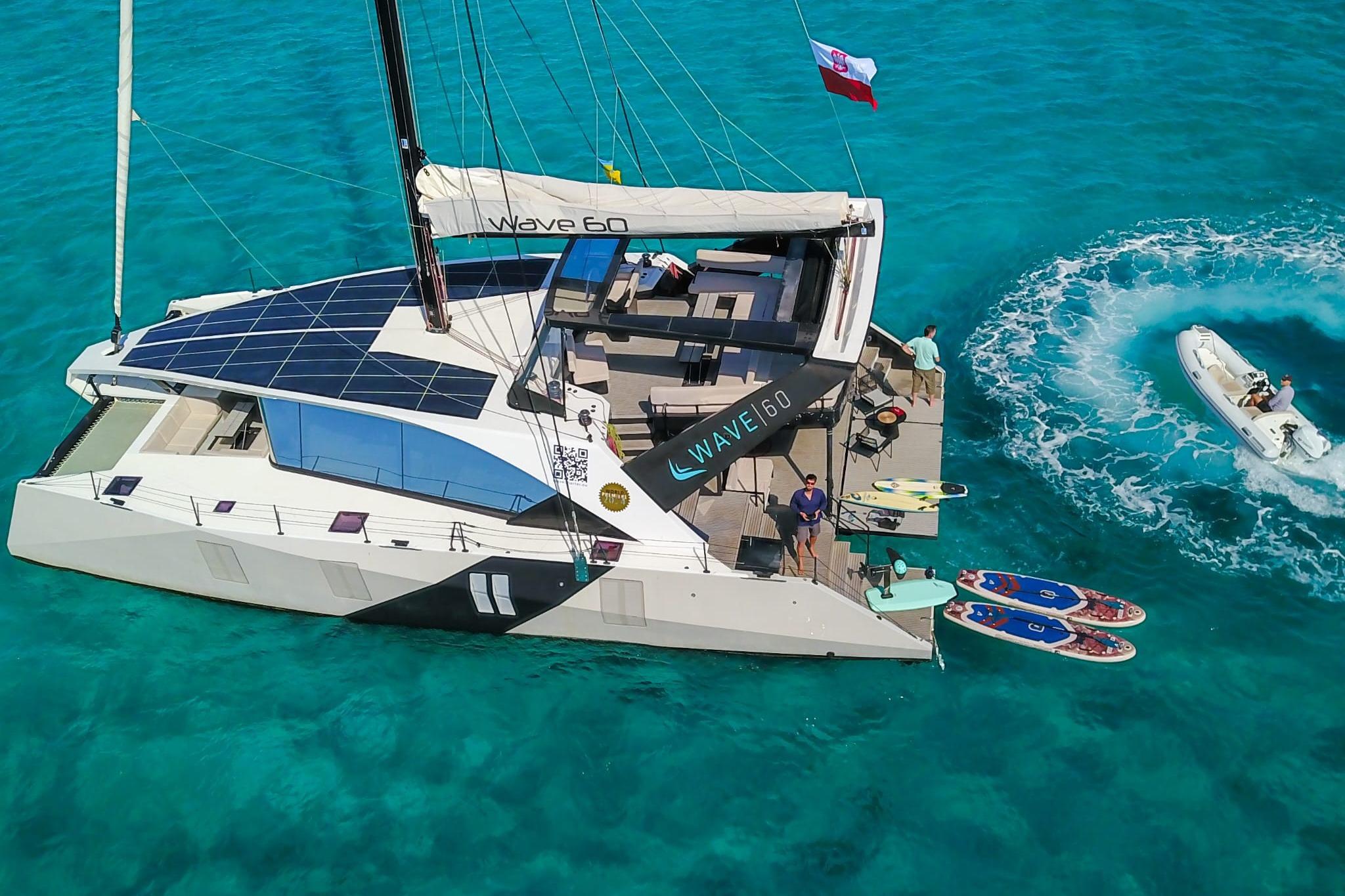 2024 Wave60 catamaran with solar panels, paddleboards, and a small boat in turquoise waters.