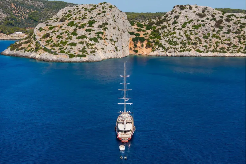 The Langley Yacht Photos Pics Aegean Yacht 3 Mast Schooner, 2009, anchored in a serene blue bay with rocky cliffs.