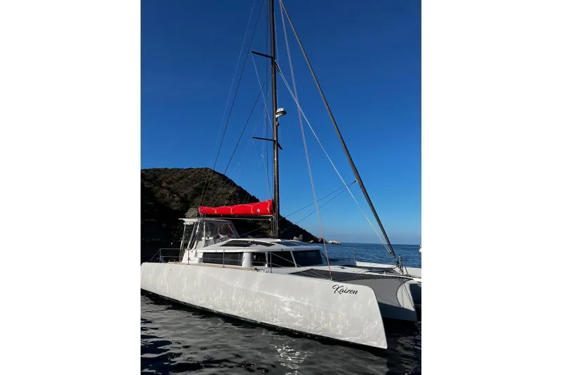 Keizan Yacht Photos Pics 2023 NEEL 43 trimaran sailing near rocky coastline under clear blue sky.