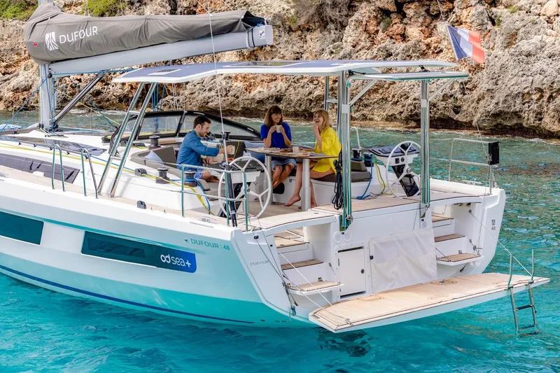  Yacht Photos Pics 2025 Dufour 48 yacht with people dining, anchored in clear turquoise water near rocky shore.