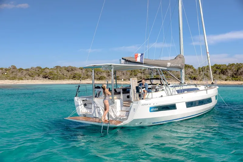  Yacht Photos Pics 2025 Dufour 48 yacht anchored in clear turquoise waters near a sandy beach.