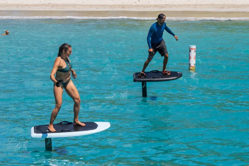 Jewel Yacht Photos Pics Two people enjoying e-foiling on clear turquoise water near a sandy beach.