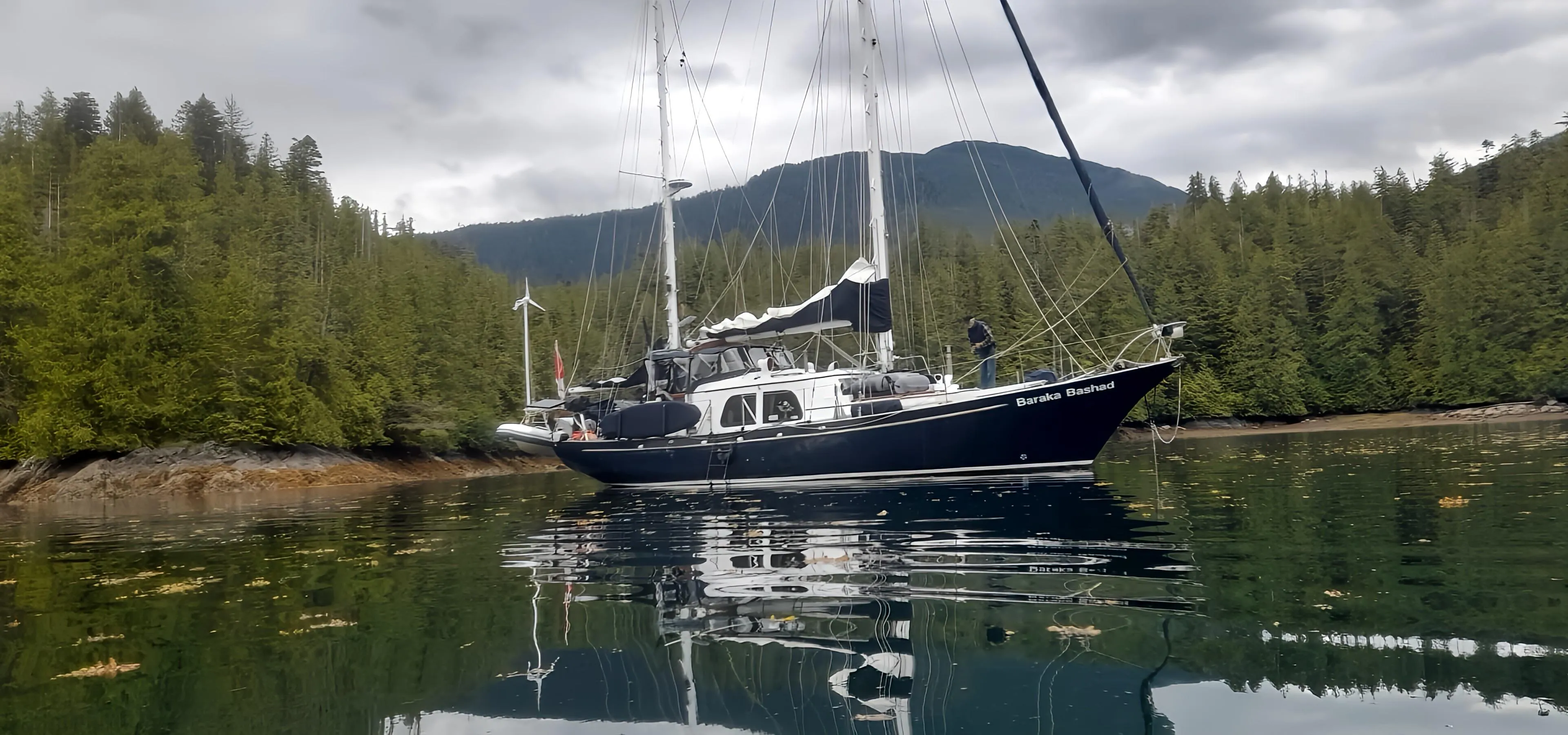 1969 Custom Rhodes Motorsailer Steel Ketch anchored in serene forested bay.