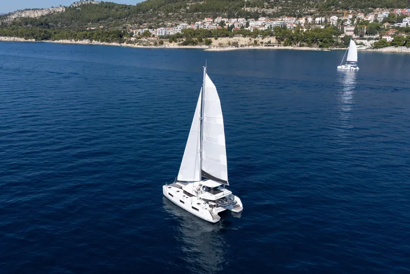 Aura Yacht Photos Pics 2024 Lagoon 46 catamaran sailing on calm blue waters near a scenic coastline.