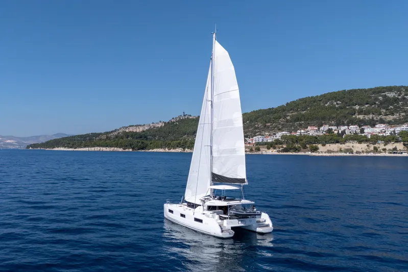 Aura Yacht Photos Pics 2024 Lagoon 46 catamaran sailing near a scenic coastline under clear blue skies.