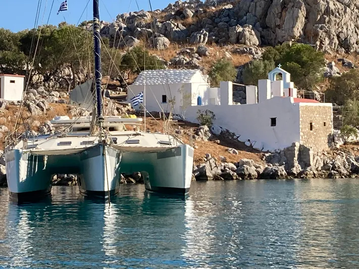 Conan Yacht Photos Pics 1988 Joubert Custom Trimaran anchored near a rocky coastline with white buildings.