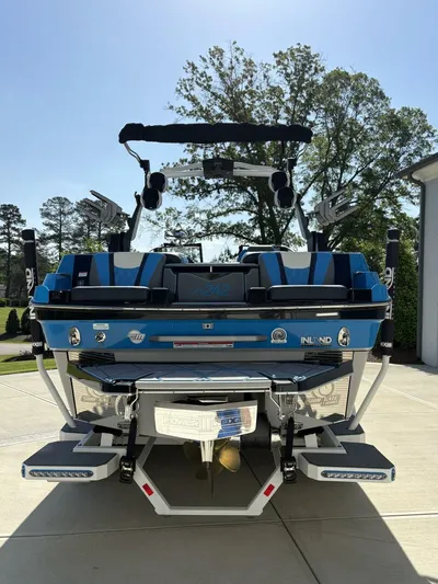  Yacht Photos Pics 2024 Malibu M242 boat, blue exterior, parked on driveway, rear view with trees in background.