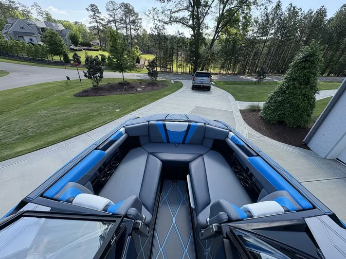  Yacht Photos Pics 2024 Malibu M242 boat with spacious seating, parked on a driveway surrounded by greenery.