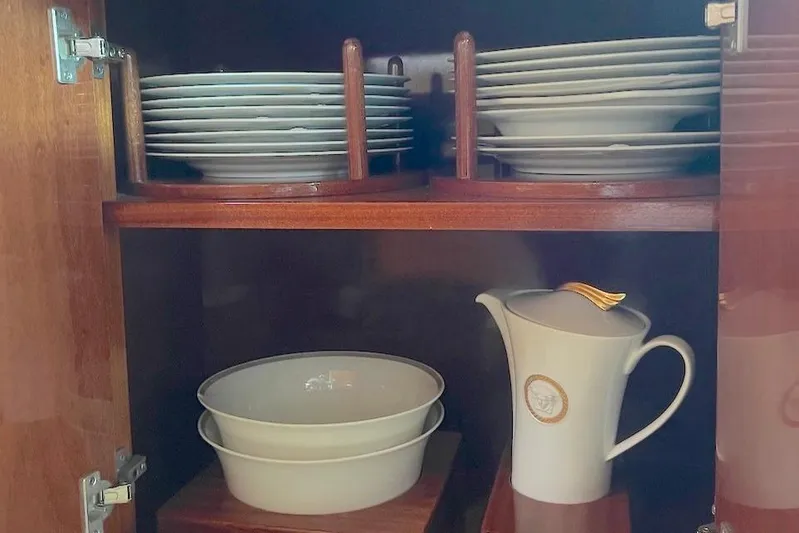 Curiosity Yacht Photos Pics Wooden cabinet with neatly stacked white plates, bowls, and a decorative pitcher.
