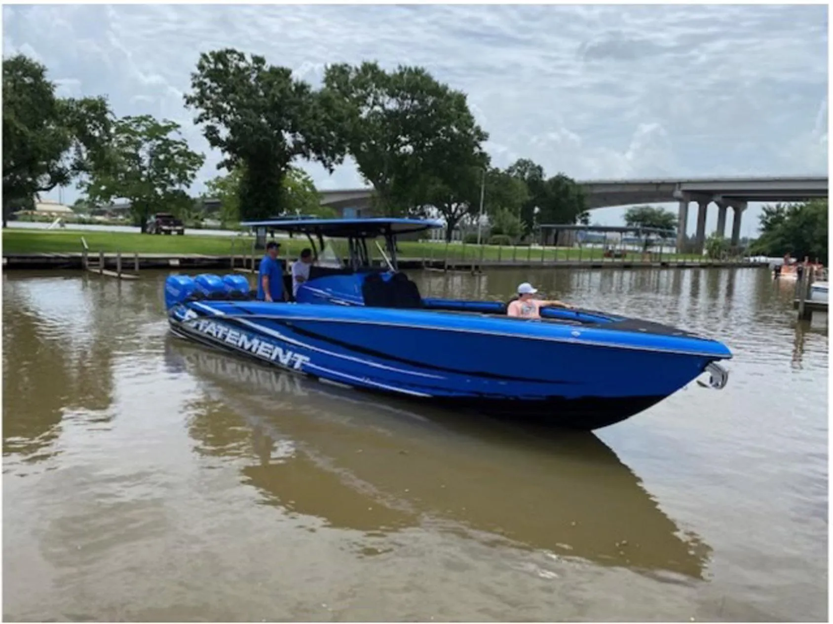 Blue 2021 Statement 380 Open boat on calm water near a bridge and trees.