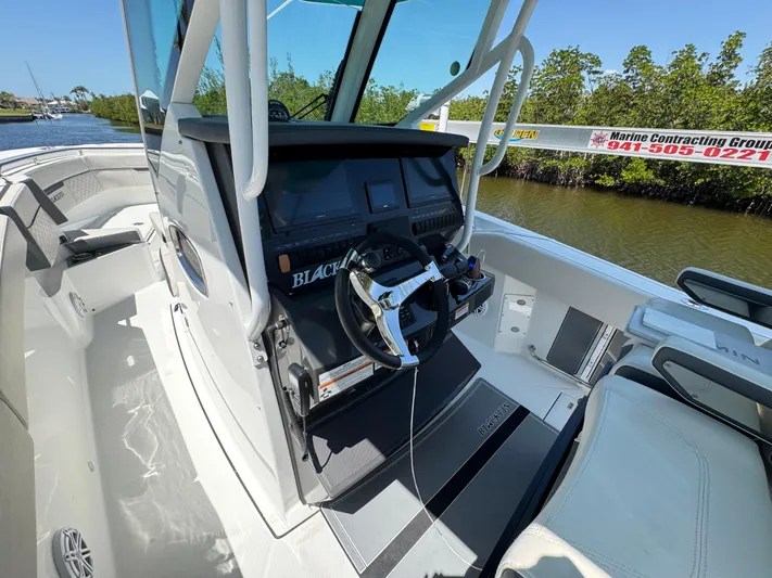  Yacht Photos Pics 2021 Blackfin 272 CC boat cockpit with steering wheel and control panel, docked near lush greenery.