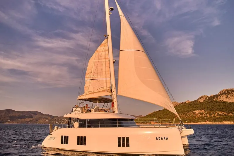Adara Yacht Photos Pics 2020 Sunreef 50 catamaran sailing near scenic coastline at sunset.