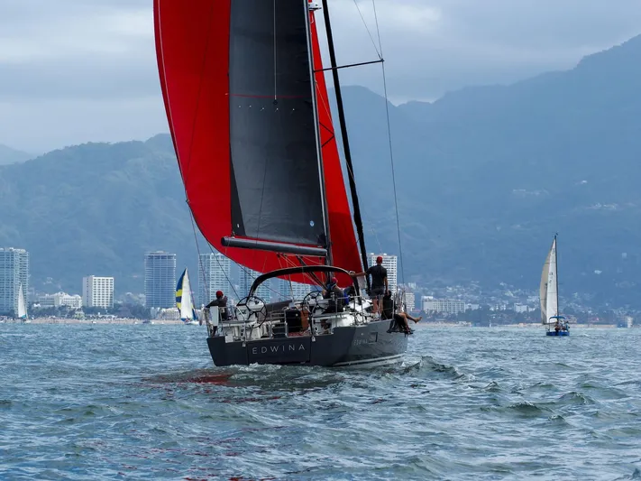 Edwina Yacht Photos Pics 2018 Jeanneau 54 sailboat with red sails on the water, cityscape and mountains in background.