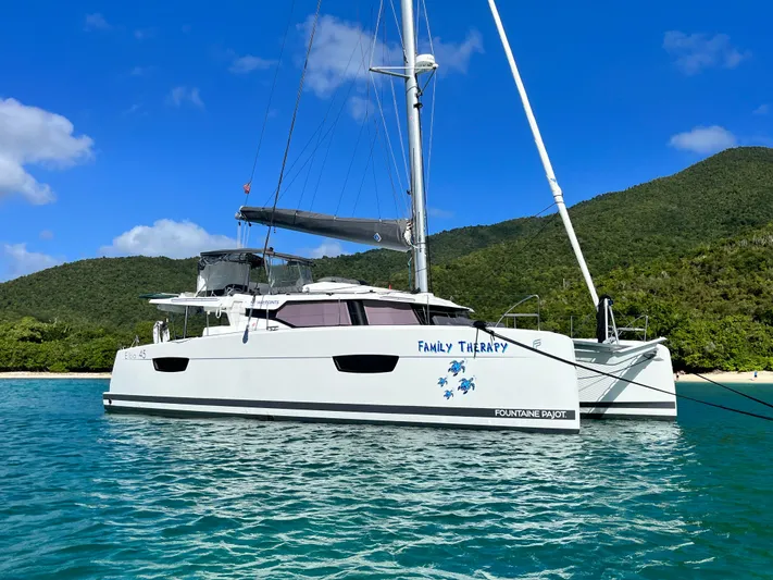 Family Therapy Yacht Photos Pics 2023 Fountaine Pajot Elba 45 catamaran anchored in clear blue water near lush green hills.