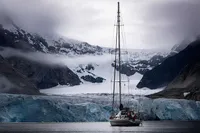 Sailboat in icy waters with glacier and snow-covered mountains in the background, under cloudy skies.