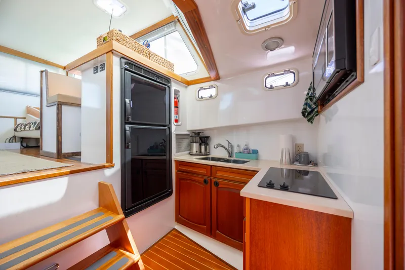 Tomahawk Yacht Photos Pics Compact boat kitchen with wooden cabinets, stove, and refrigerator, featuring natural light from windows.