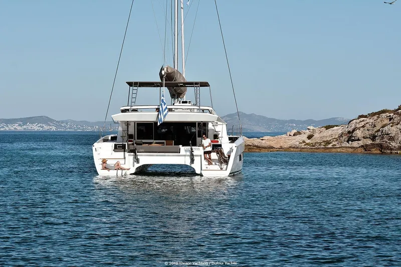  Yacht Photos Pics 2020 Dufour Catamarans 48 sailing near rocky coastline under clear blue sky.