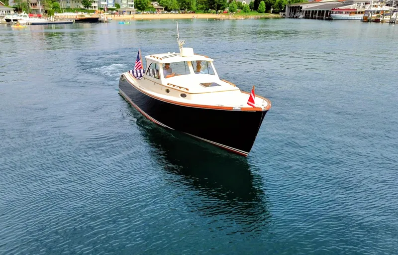 Luna Yacht Photos Pics Hinckley Picnic Boat 2000 cruising on a serene lake near a bustling shoreline.