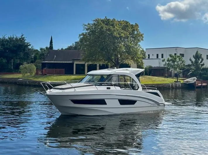  Yacht Photos Pics 2025 Beneteau Antares 9 boat on a calm river, surrounded by greenery and houses.
