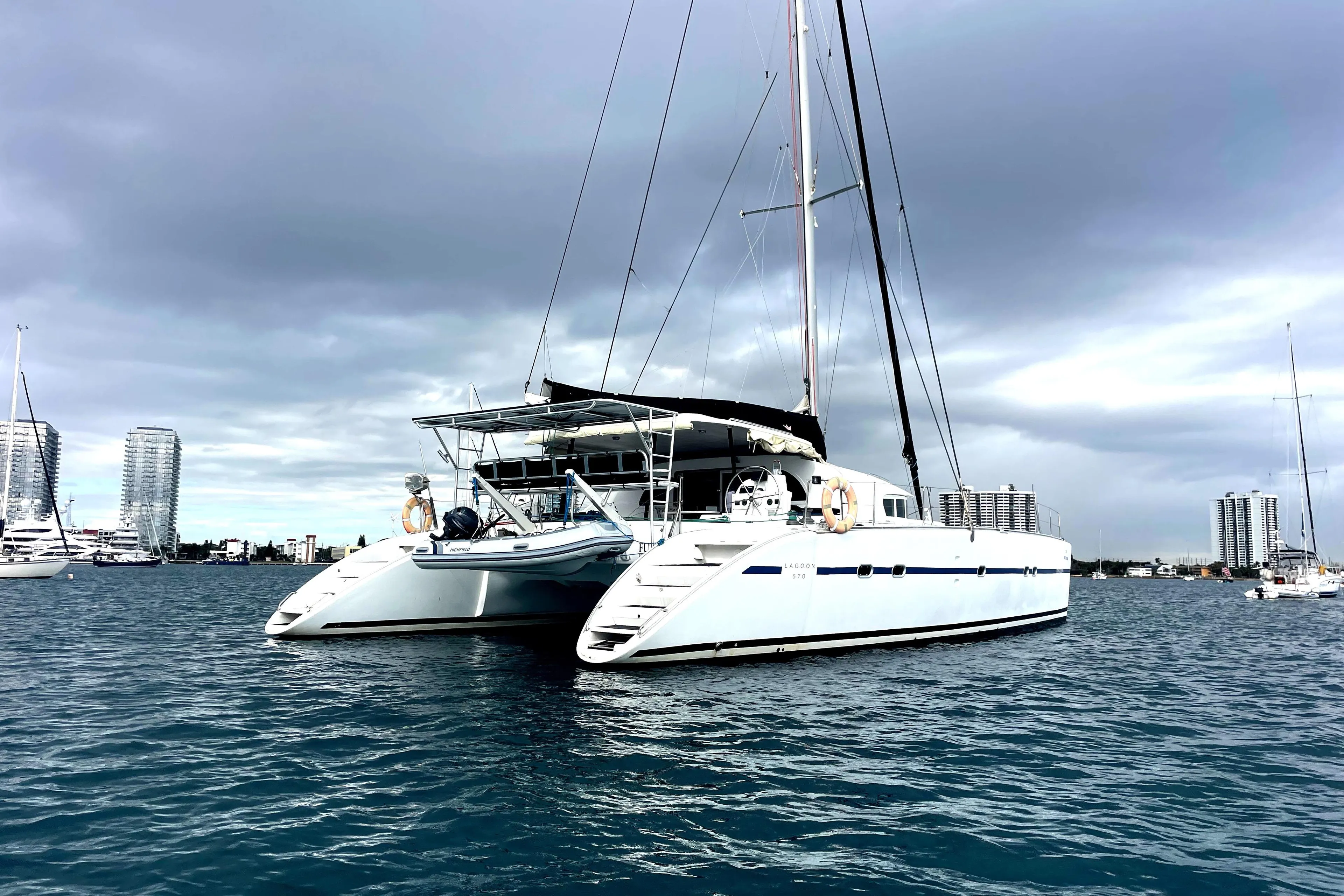 2004 Lagoon 570 catamaran anchored in a marina with city skyline backdrop.