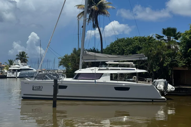 Ten:10 Yacht Photos Pics 2017 Fountaine Pajot SABA 50 catamaran docked by lush greenery and blue sky.
