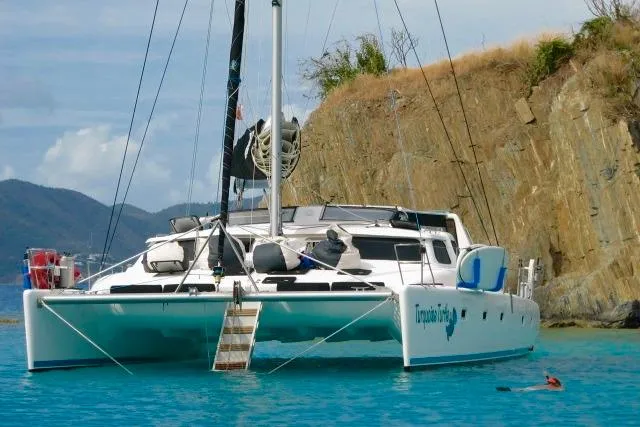 Turquoise Turtle Yacht Photos Pics Catamaran Voyage 500, 2001 model, anchored near rocky coastline in clear blue water.