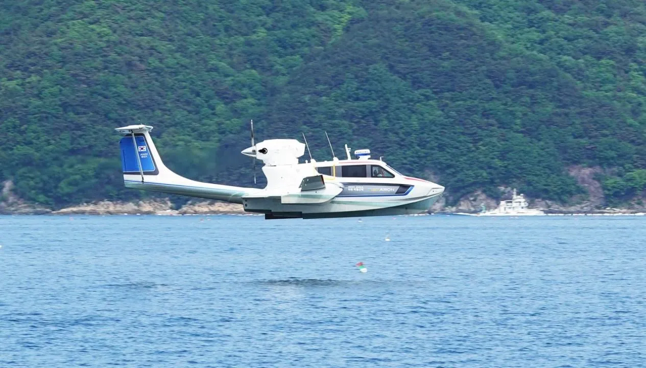 Futuristic Aron M 80 seaplane flying over water, 2025 model, with lush green hills in background.