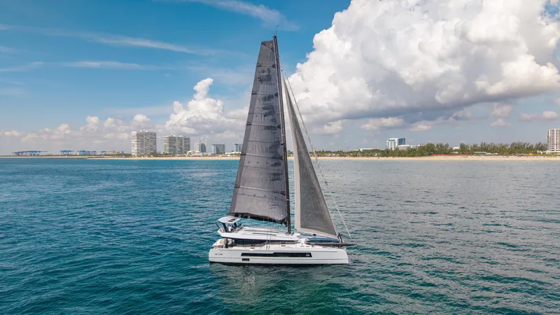 Noble House Yacht Photos Pics 2019 McConaghy MC60 sailing near a coastal city under a partly cloudy sky.