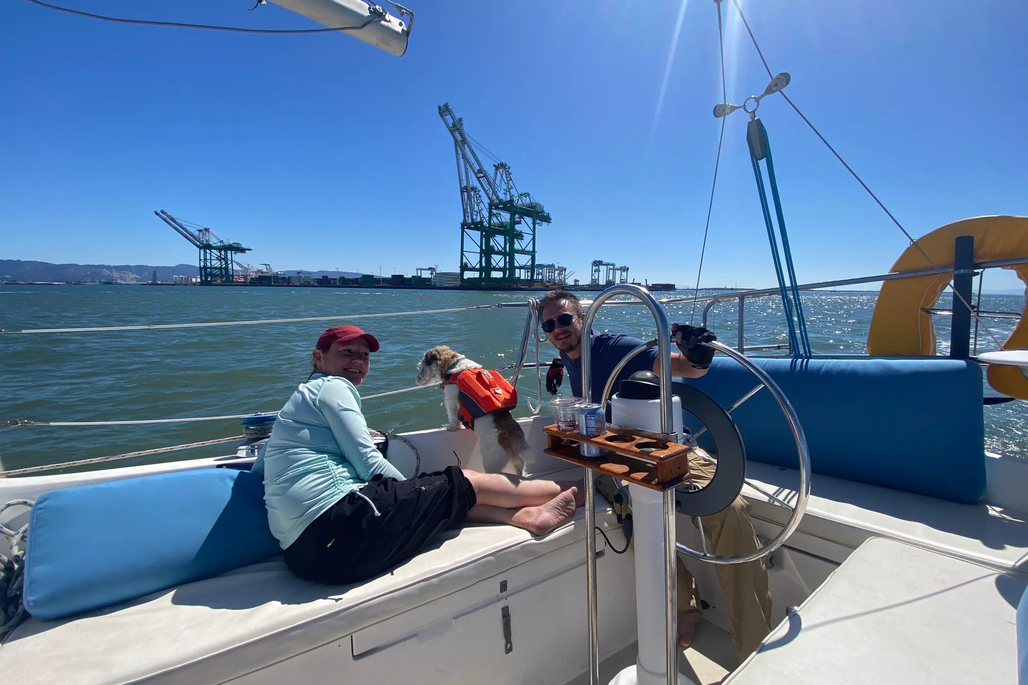 Two people and a dog on a 1980 Catalina 30 sailboat, with cranes in the background.