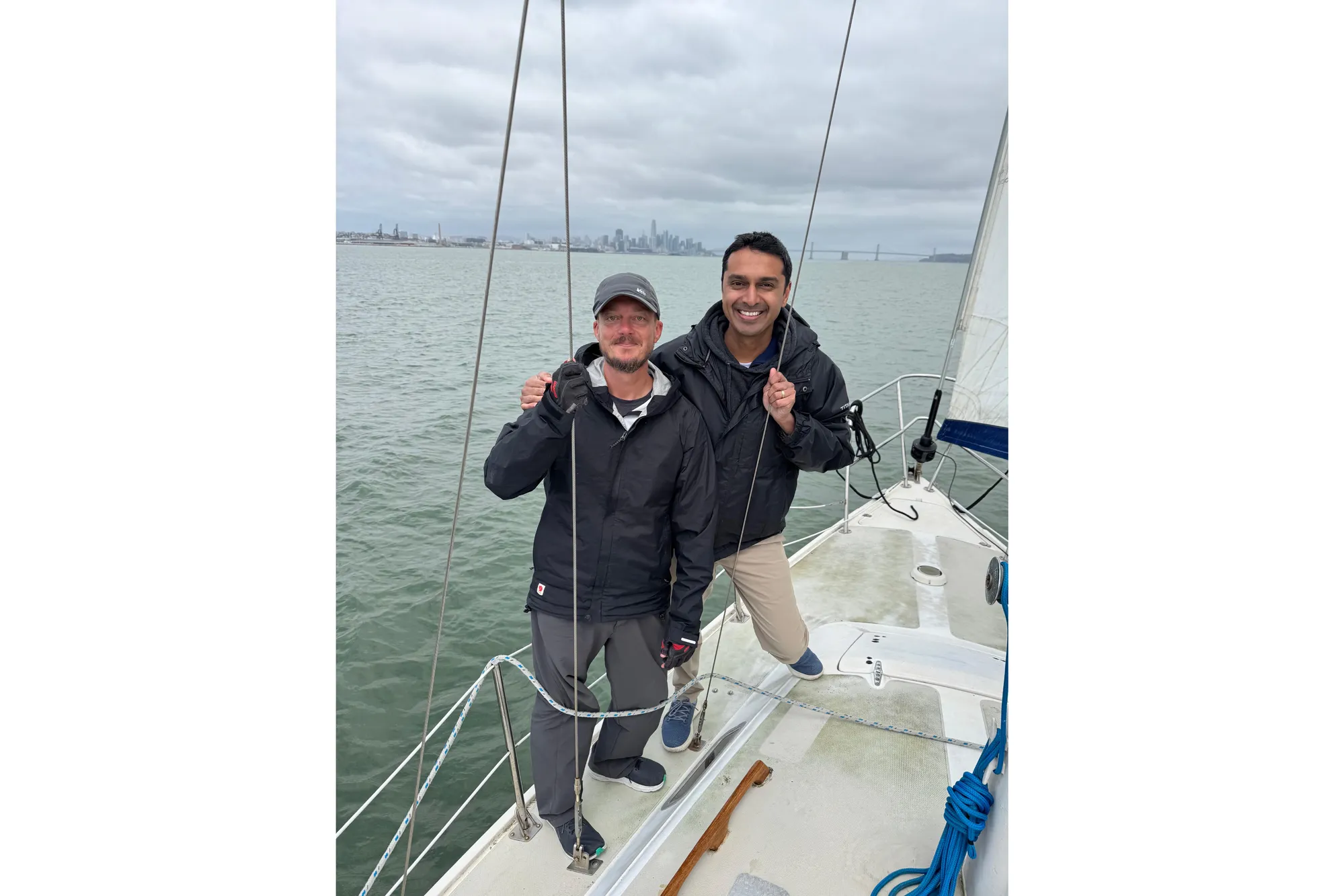 Two people smiling on a 1980 Catalina 30 sailboat, with a cityscape in the background.