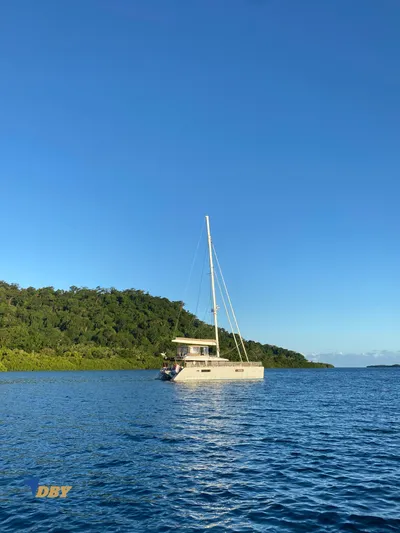 Samadhi Yacht Photos Pics 2019 Lagoon 560 S2 catamaran sailing near lush green island under clear blue sky.