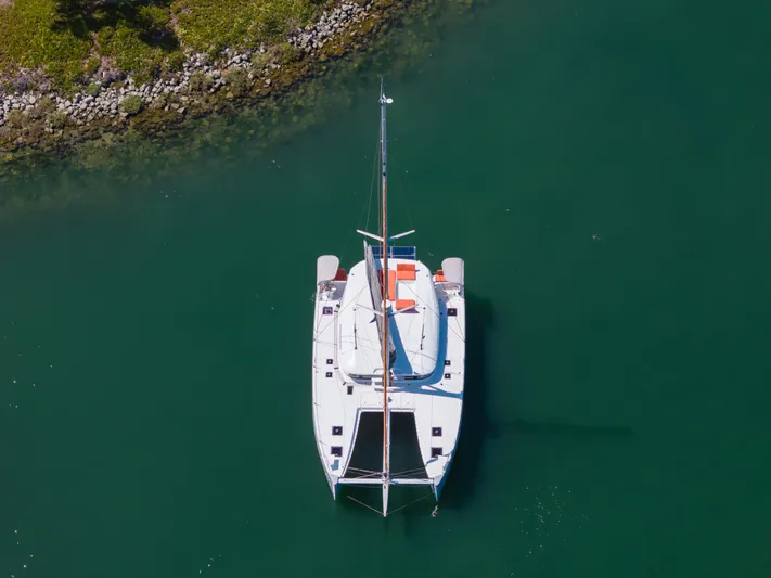 In Stock Yacht Photos Pics Aerial view of 2024 Excess 14 catamaran on calm green water near rocky shoreline.