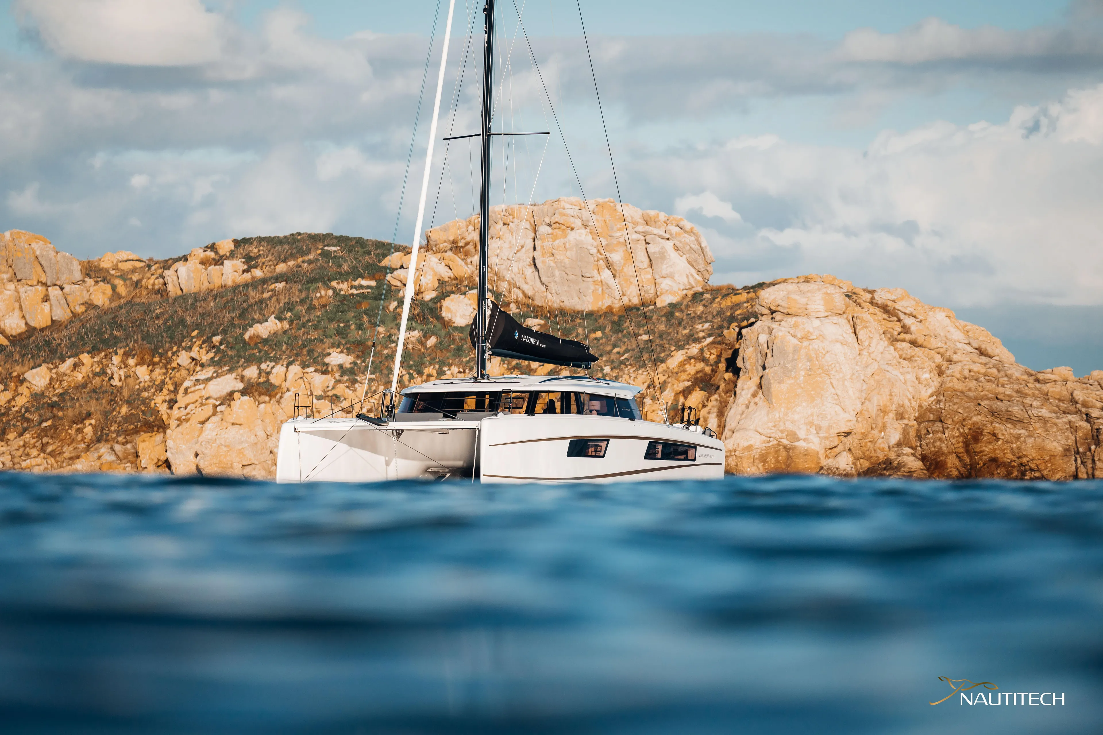 Nautitech 48 Open - Available For 2026 Delivery! Yacht Photos Pics Nautitech 48 Open 2026 catamaran sailing near rocky coastline under blue sky.