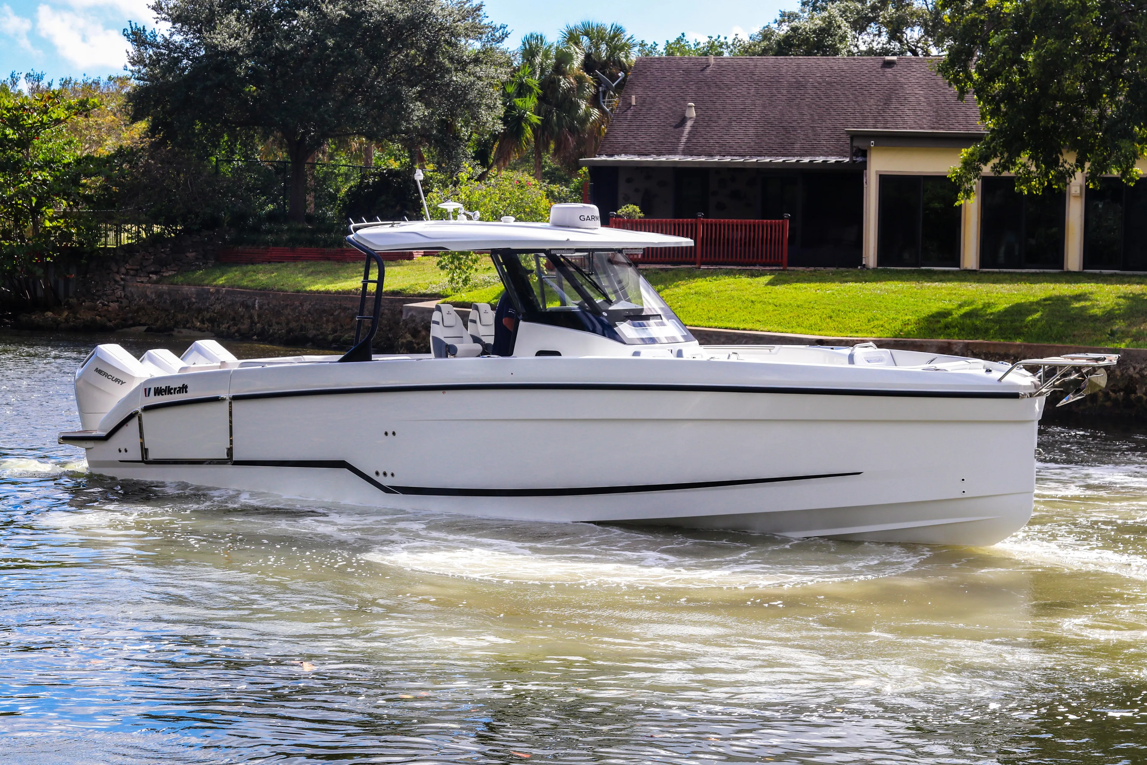 2026 Wellcraft 38 T-Top boat cruising on a sunny day near a house.