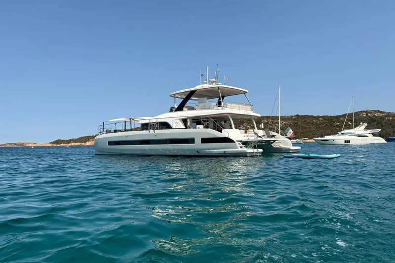 4ever VI Yacht Photos Pics 2022 Lagoon 67 yacht anchored in clear blue waters, with scenic coastal backdrop.