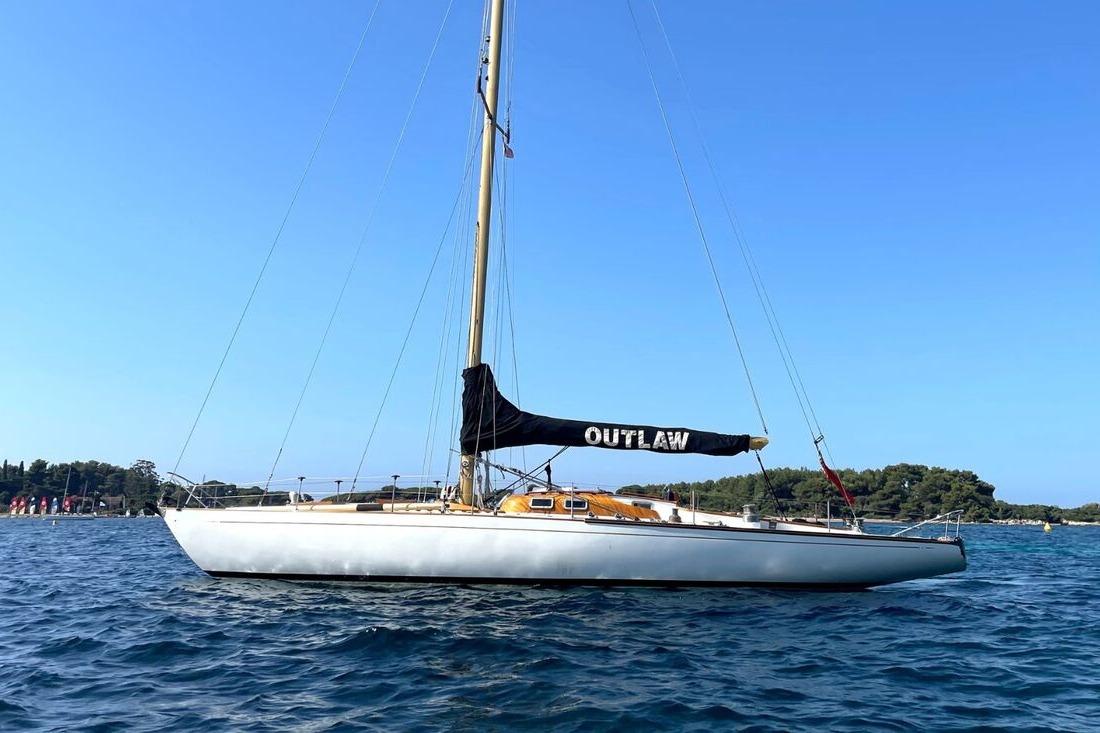 1963 Souters of Cowes sailboat "Outlaw" on calm blue water, clear sky background.