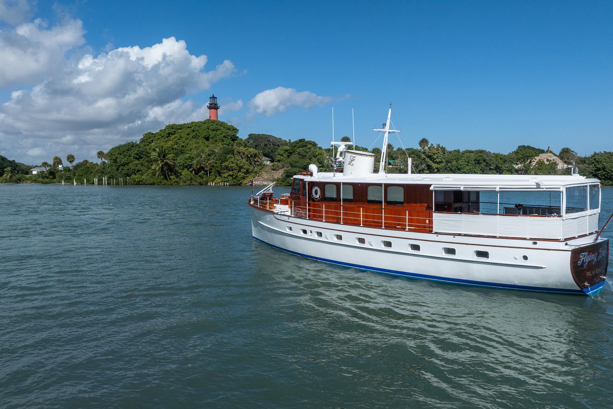 Vintage 1937 Trumpy 58 yacht cruising near a scenic lighthouse and lush greenery.