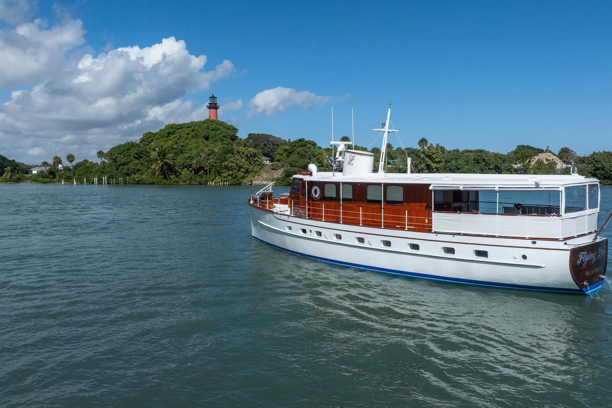 Vintage 1937 Trumpy 58 yacht cruising near a scenic lighthouse and lush greenery.