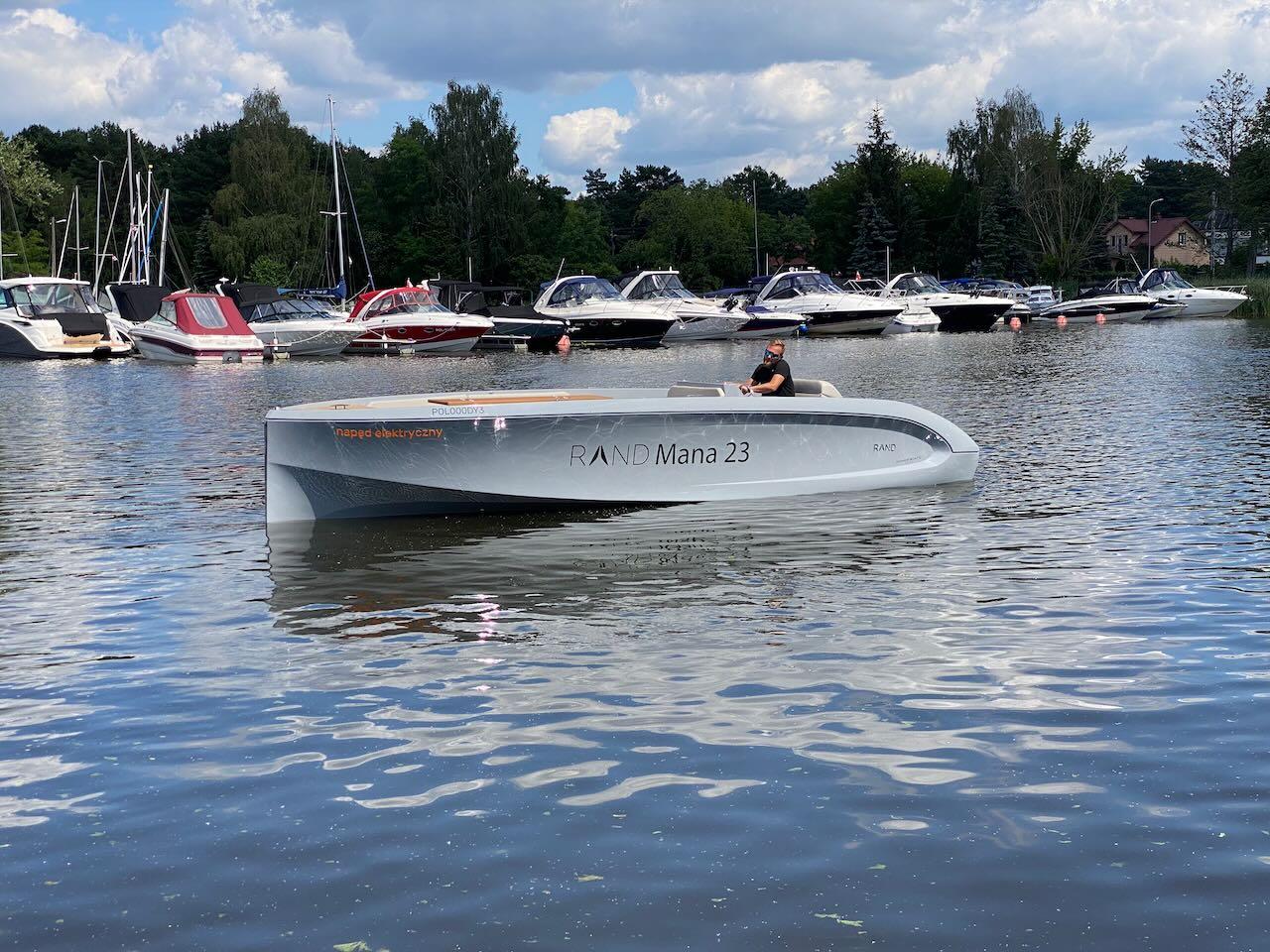 2021 Rand Mana 23 boat on a calm lake with other boats docked in the background.