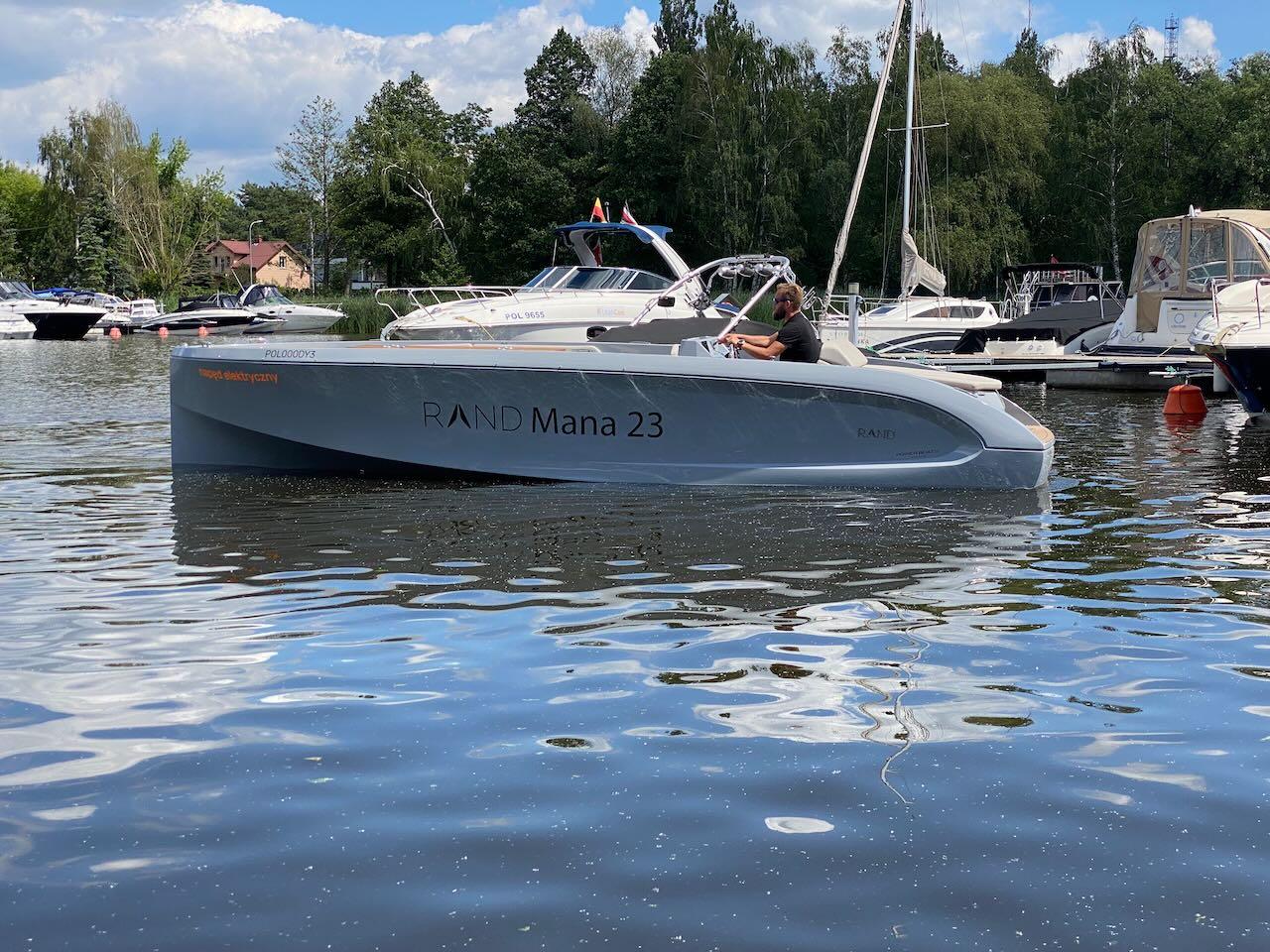 Modern 2021 Rand Mana 23 boat cruising on a serene lake with other boats docked nearby.