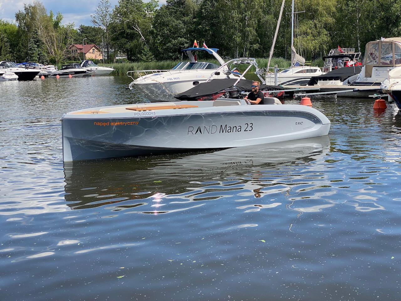2021 Rand Mana 23 electric boat on a calm lake near a marina.