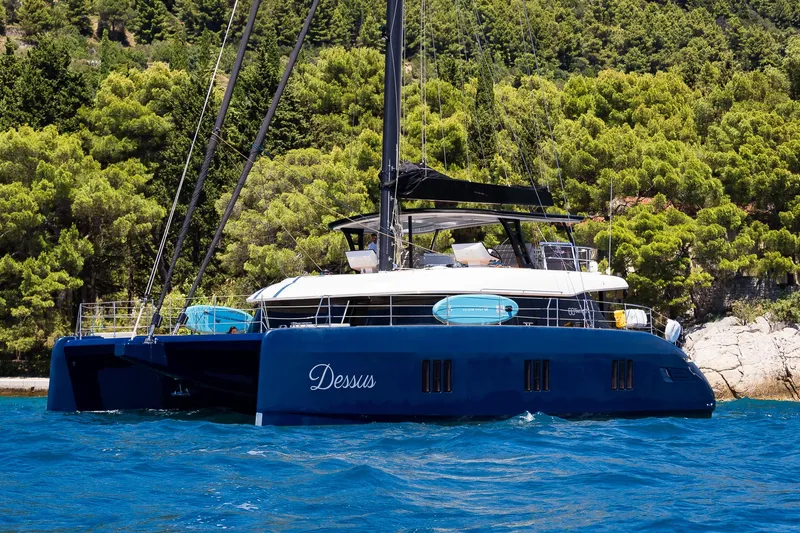 Dessus Yacht Photos Pics Luxurious 2022 Sunreef 60 catamaran sailing near a lush, forested coastline.