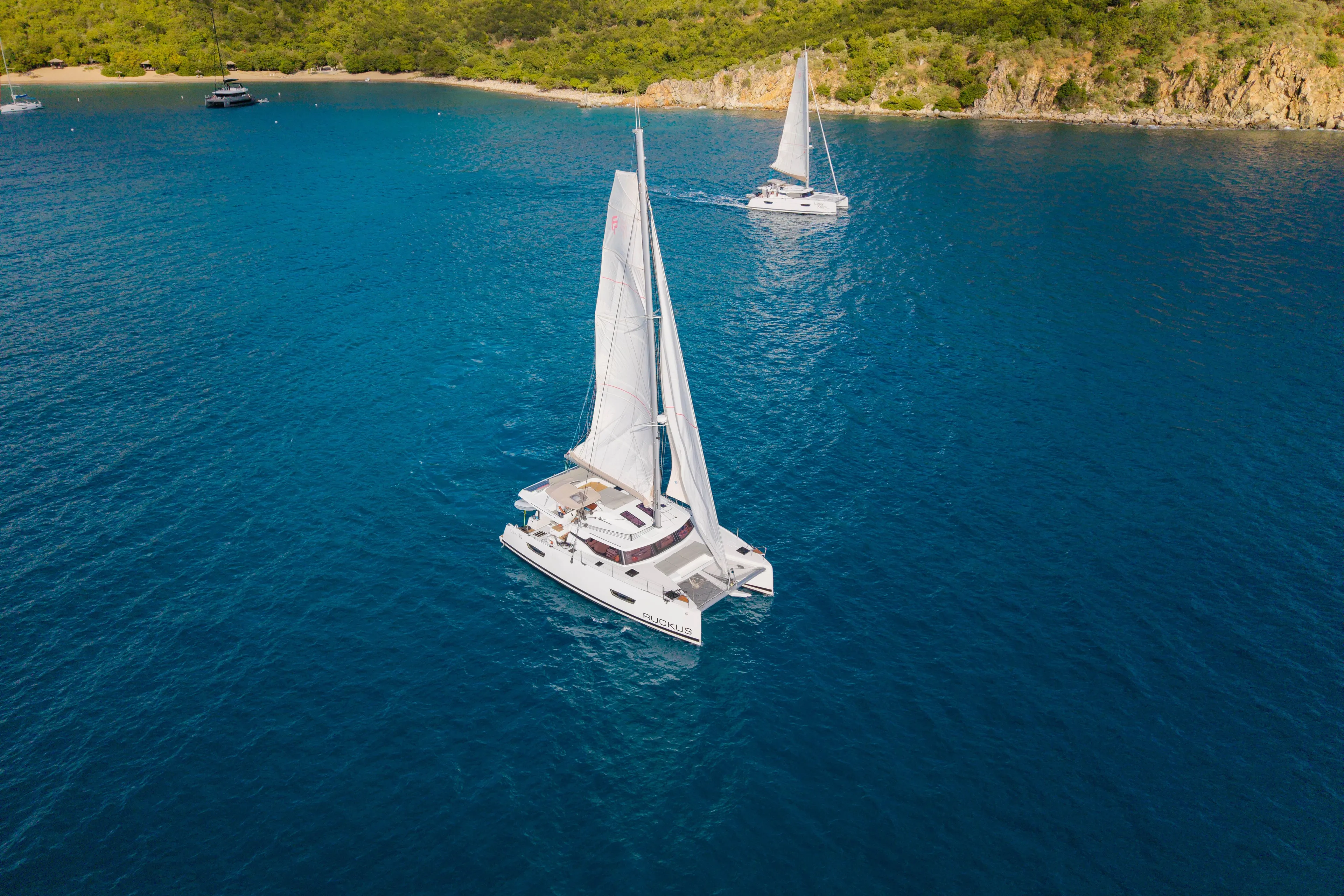 Ruckus Yacht Photos Pics 2021 Fountaine Pajot Elba 45 Catamaran sailing in clear blue waters near a lush coastline.