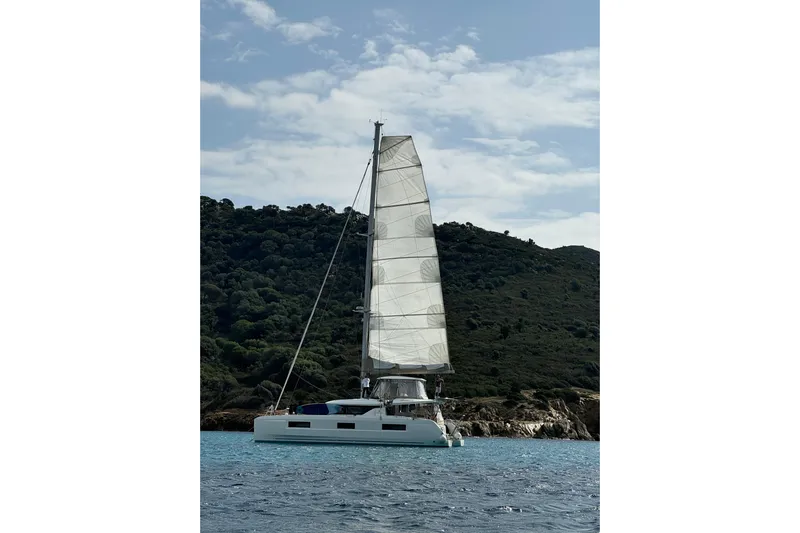  Yacht Photos Pics 2022 Lagoon 46 catamaran sailing near a lush, hilly coastline under a partly cloudy sky.