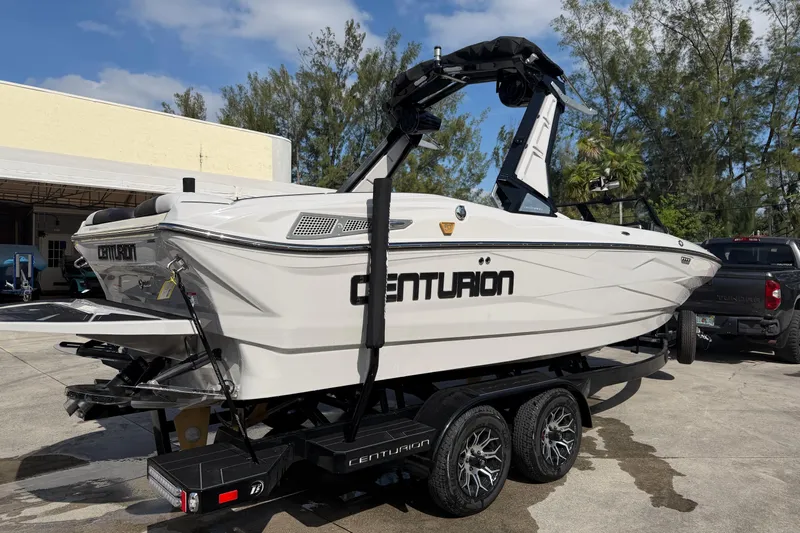  Yacht Photos Pics 2026 Centurion Nv233 boat on trailer, parked outdoors, with trees and building in background.