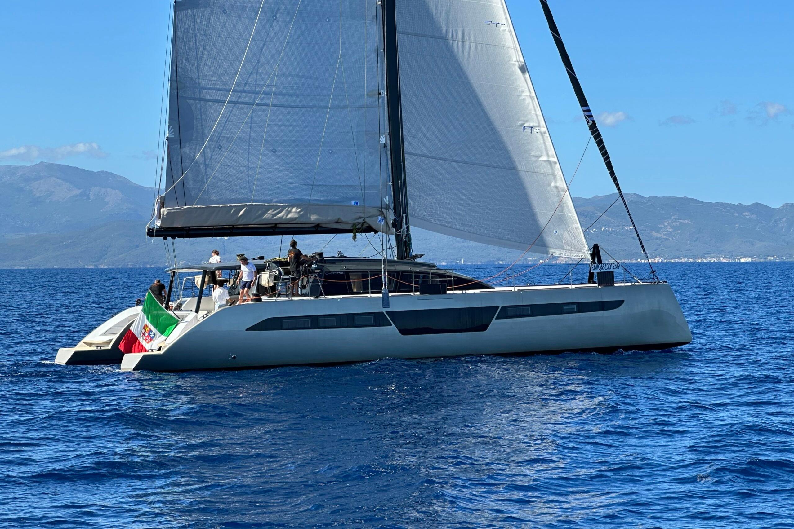 2026 Comar C-CAT 65 catamaran sailing on blue ocean with mountainous backdrop.